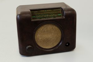 A brown Bakelite radio.  There is a panel at the top showing the two radio bands with the stations marked on them along their length.  The round speaker is in the middle, covered with mesh.  The tuning handle is on the bottom left hand side and the on/off switch is on the right hand side.