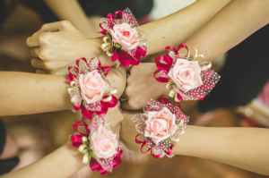 Three pairs of bridesmaids' hands meeting together in a circle.  They are wearing bracelets made of pale pink roses and bright pink ribbons.