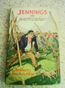 The cover of a book called Jennings in particular, showing a teacher angrily chasing a piglet through a broken fence, in front of a crowd of laughing schoolboys, all wearing school caps. 
