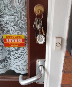 A close-up of the door handle, door bell and keyhole with a bunch of keys hanging from it on a Manchester City football club key ring.  There is a sticker in the window saying "Burglars beware, our property is protected".
