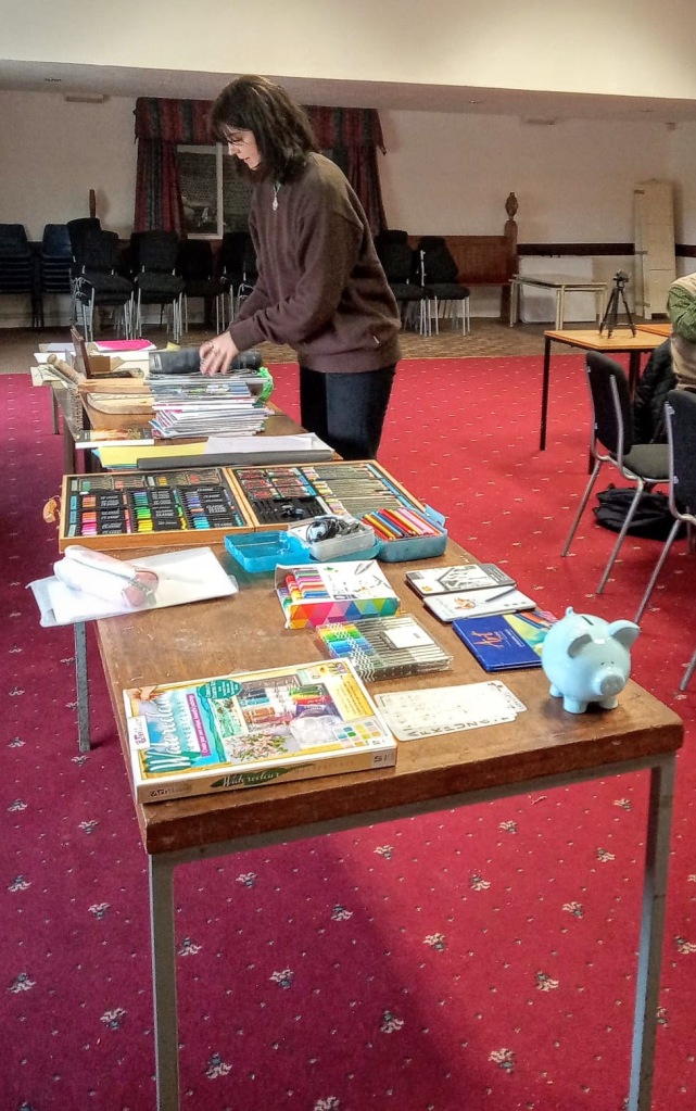 Georgia is setting out an array of pens, pencils, coloured paper and other art materials on two large tables.